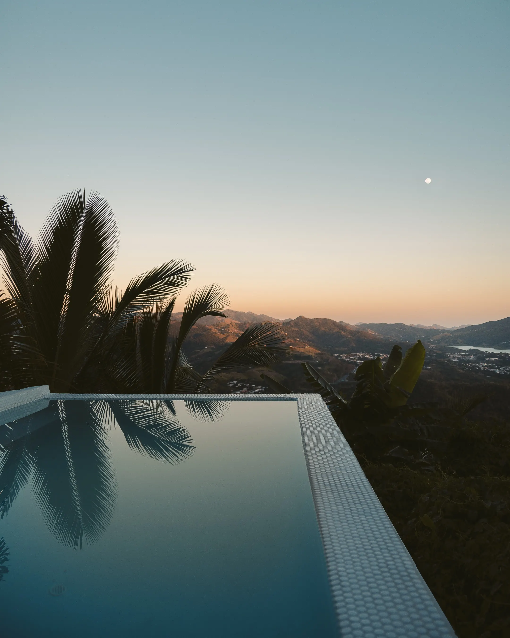 A beautifull morning in the mountains. The moon is still in the sky while the lower part of the sky is turning yellow. A pool and palmtrees are in the forground and a town is down in a vally in the background.